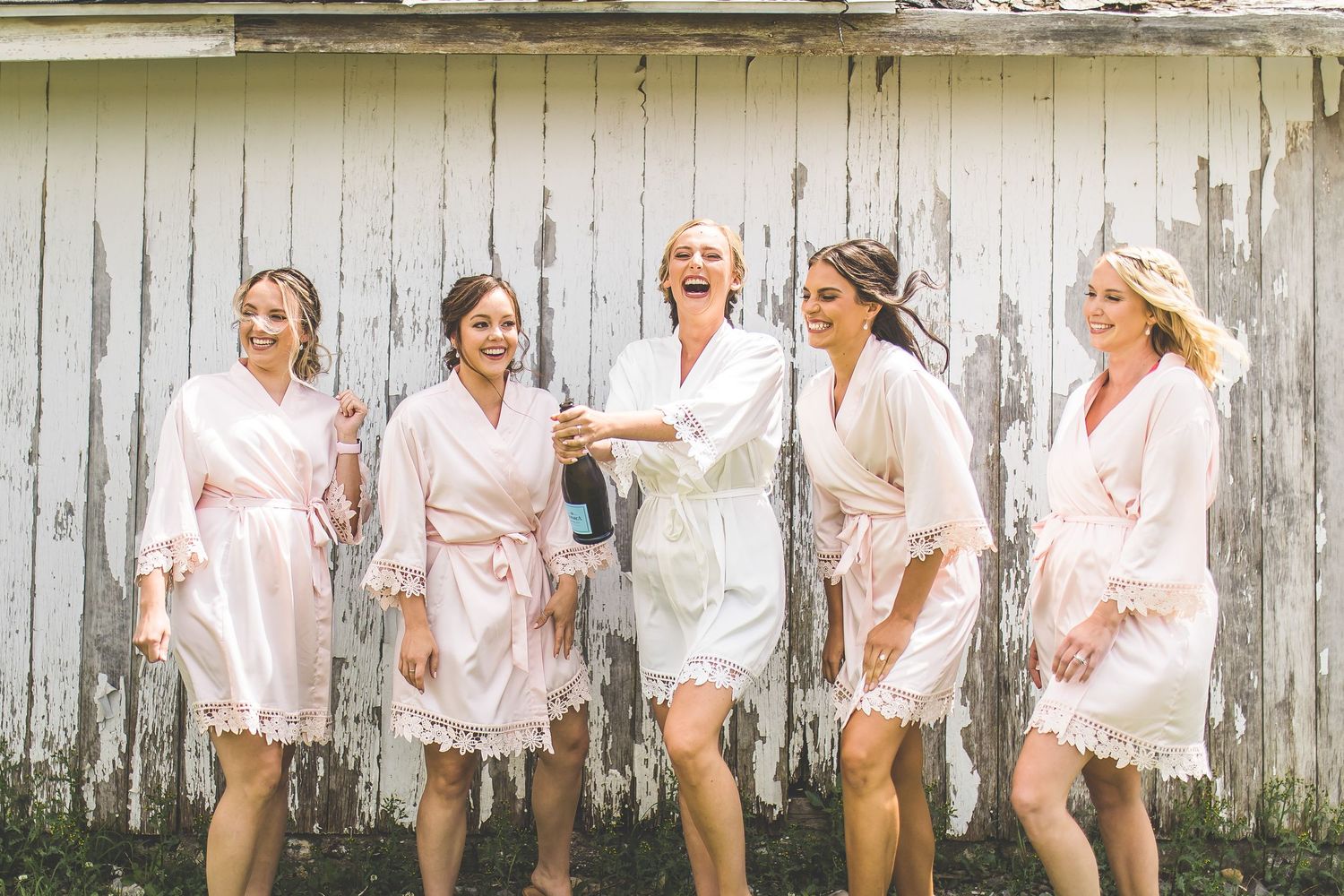 Bridesmaids in white robes laughing together against weathered wooden wall.