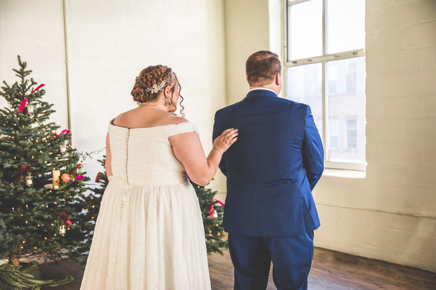 Couple in wedding attire standing together near decorated Christmas trees in bright room.