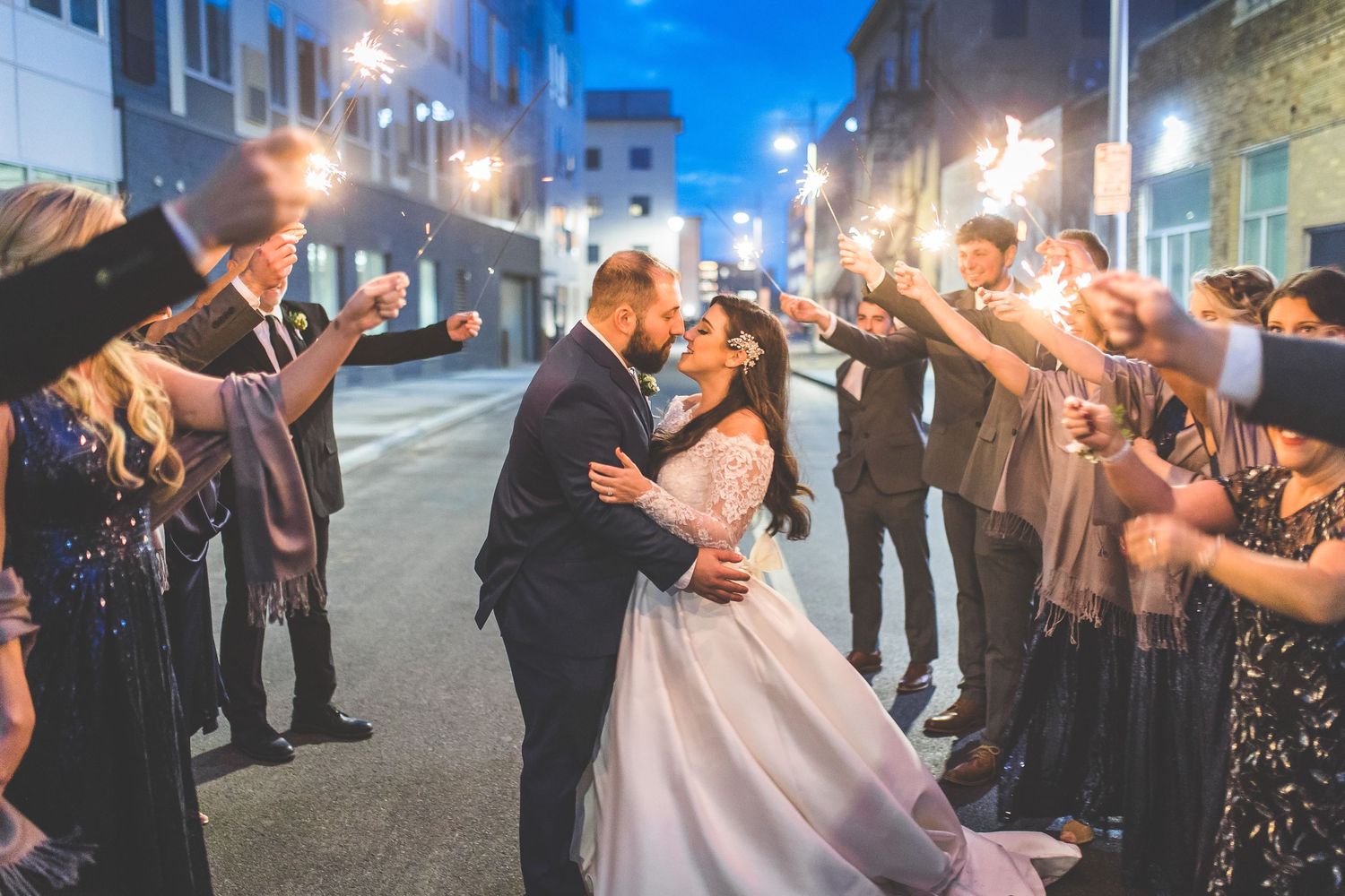 Newlyweds share romantic kiss on city street at night surrounded by sparkler-holding wedding guests.