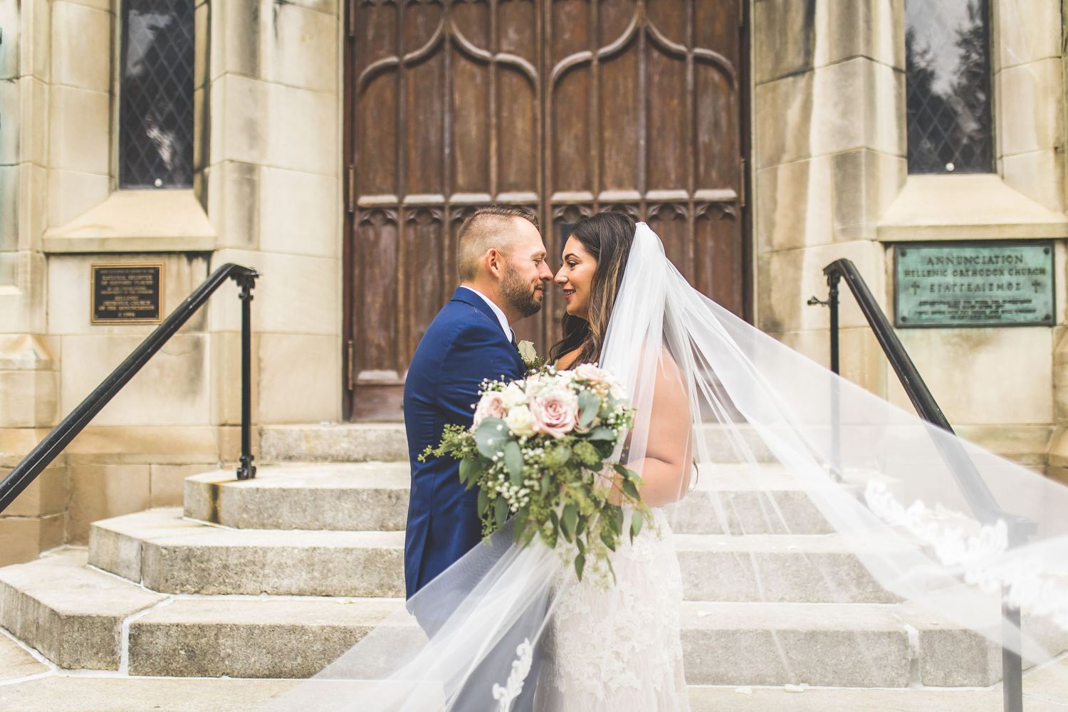 Newlyweds share intimate moment on church steps with flowing veil and floral bouquet.