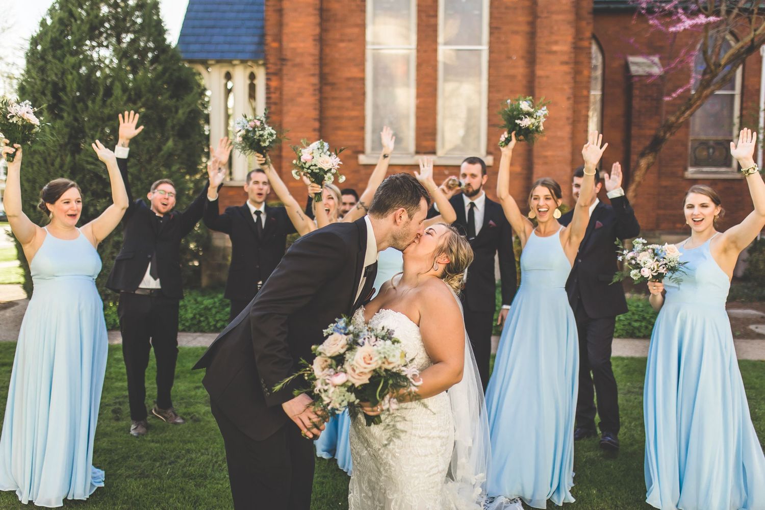 Joyful wedding party celebrates outside brick church with raised arms and bouquets.