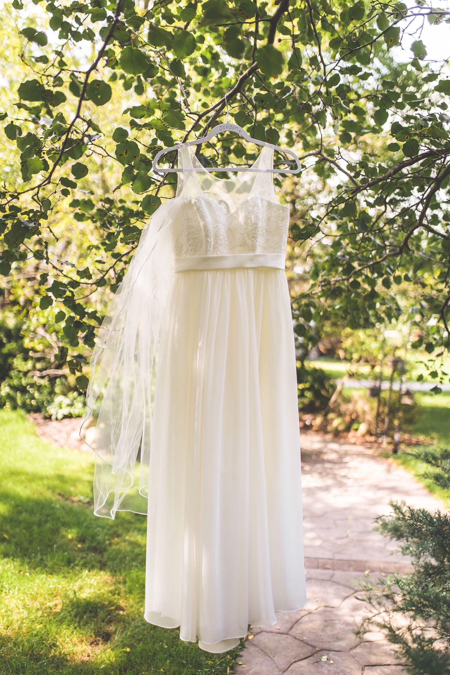 White wedding dresses hanging on hangers beneath green foliage outdoors.