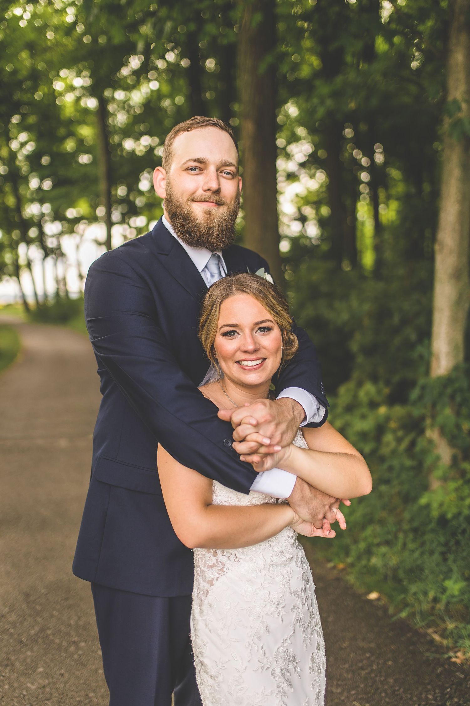Joyful wedding couple laughing and embracing on a tree-lined gravel pathway.
