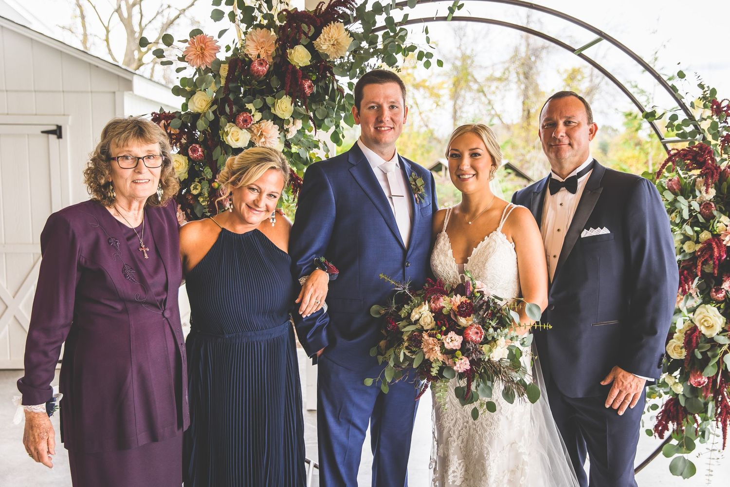 Wedding party members stand together under elegant floral arch in navy and burgundy attire.