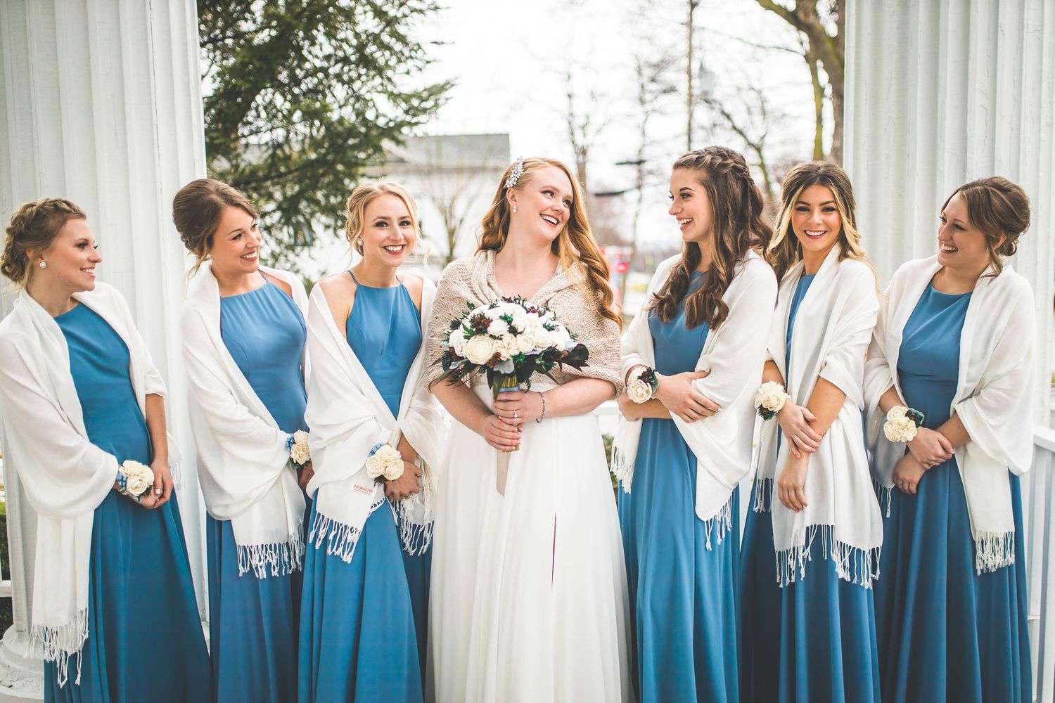 Bridesmaids in teal dresses and white shawls posing together outdoors.