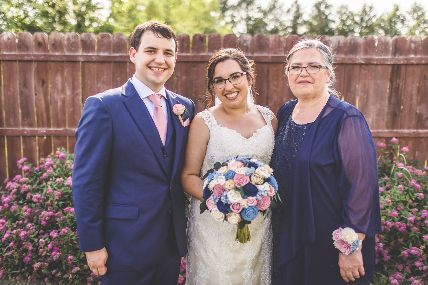 Wedding portrait of three people posing with pink and blue bouquet against wooden fence and flowers.