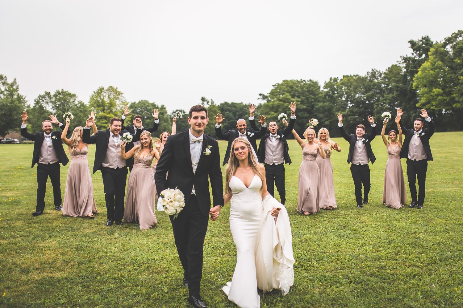 Large wedding party group photo with bridesmaids in pink and groomsmen in black on rolling lawn.