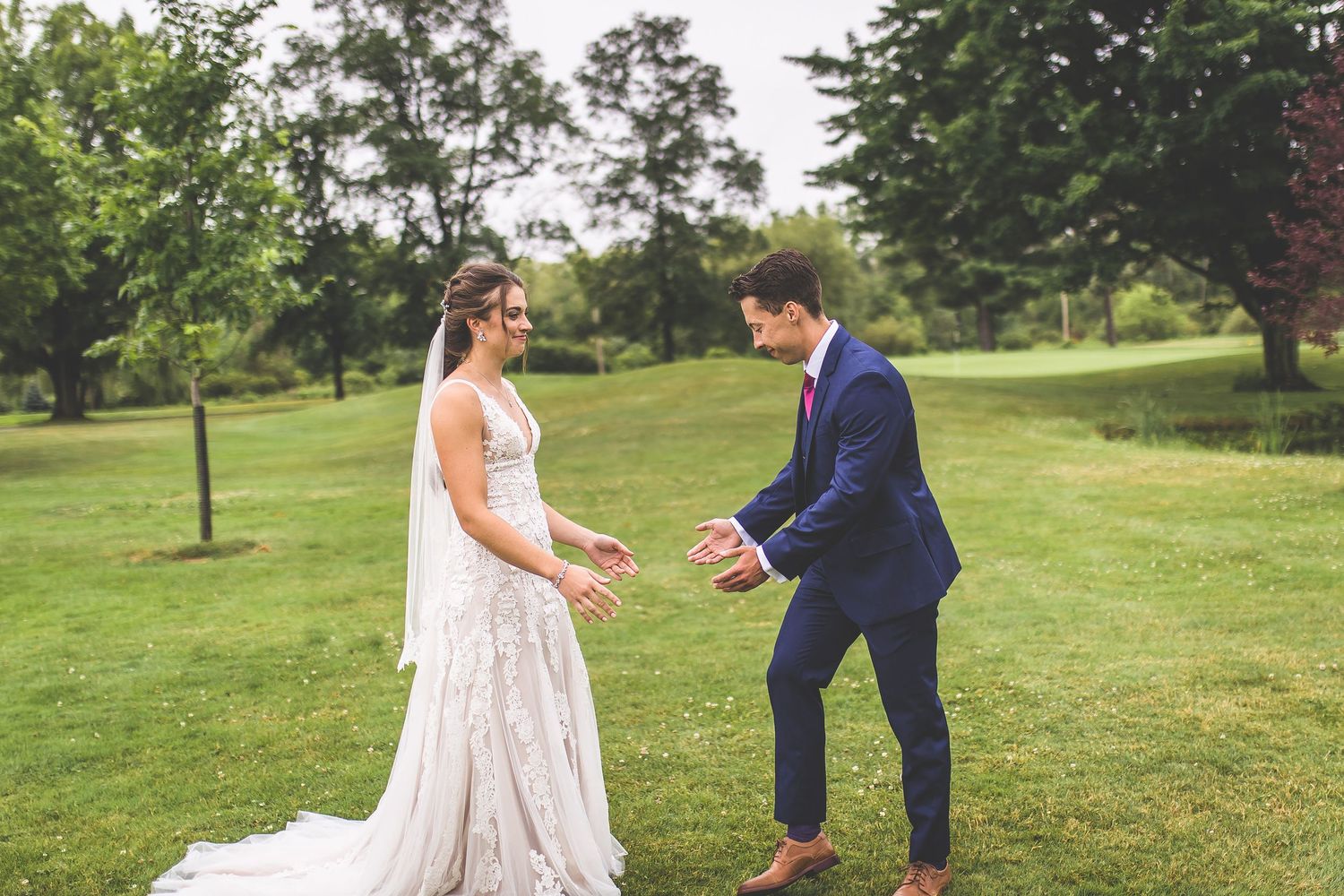 A couple shares a joyful moment during their first look on a lush green lawn with trees in the background.