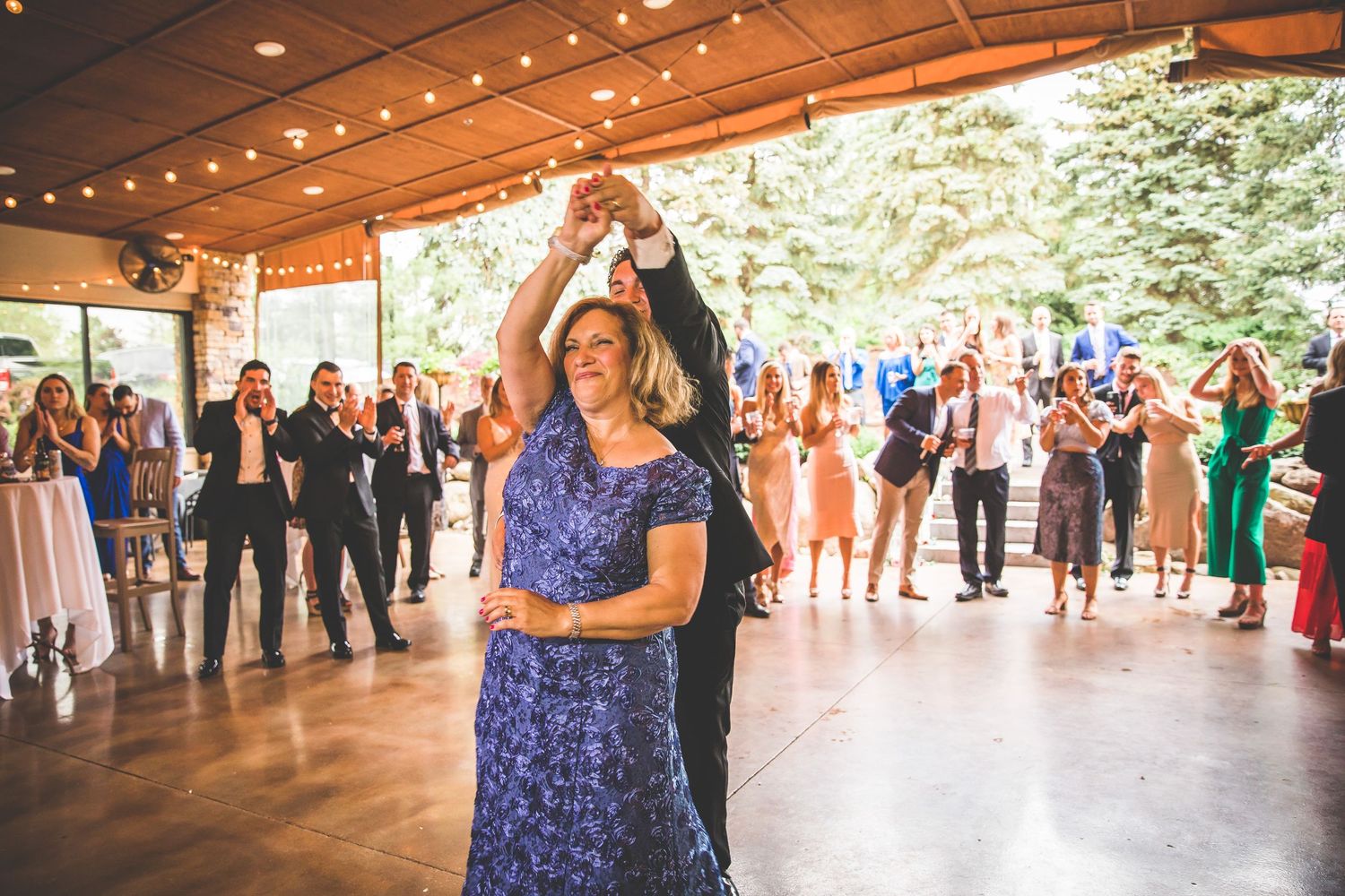 Dancing couple surrounded by guests during wedding reception in venue with forest views.