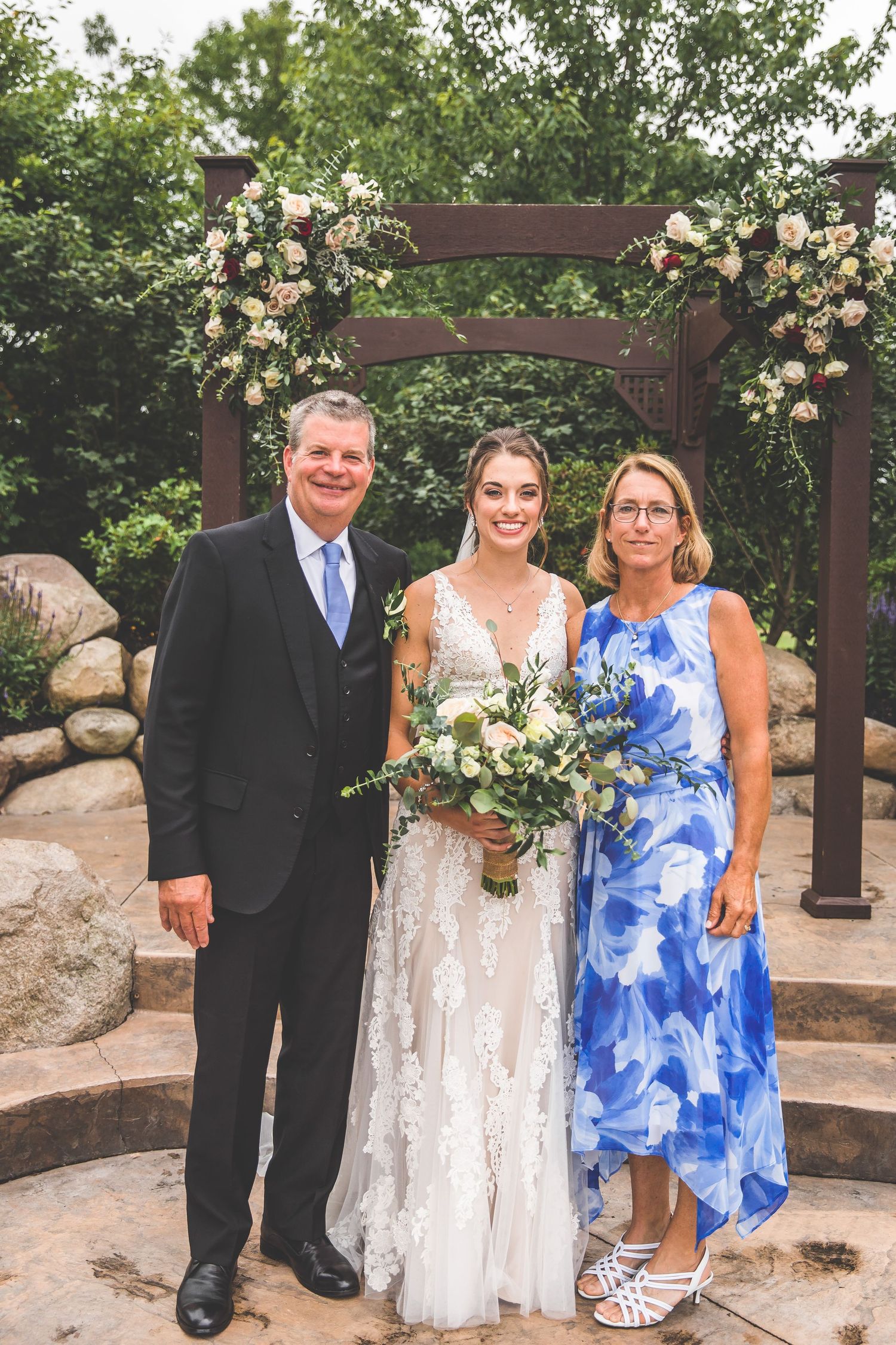 Wedding party poses for formal photos under floral-decorated wooden arch.