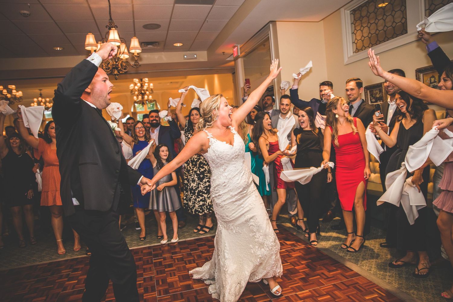 Wedding couple dances energetically while guests cheer and wave napkins in celebration.