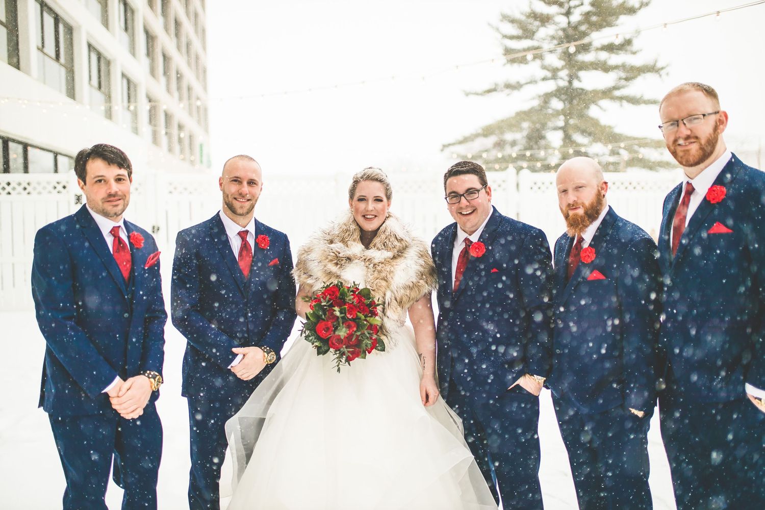 Wedding party stands in line during snowfall wearing navy suits and red accents.