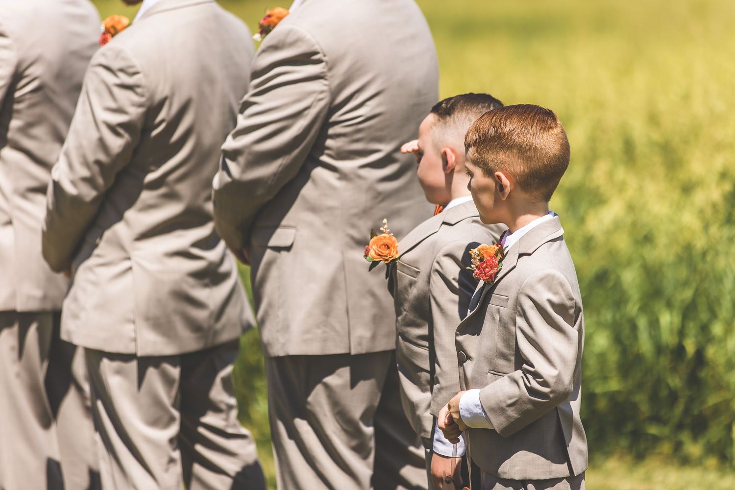 Wedding party in grey suits and bridesmaids in coral dresses gather outdoors on a sunny summer day.