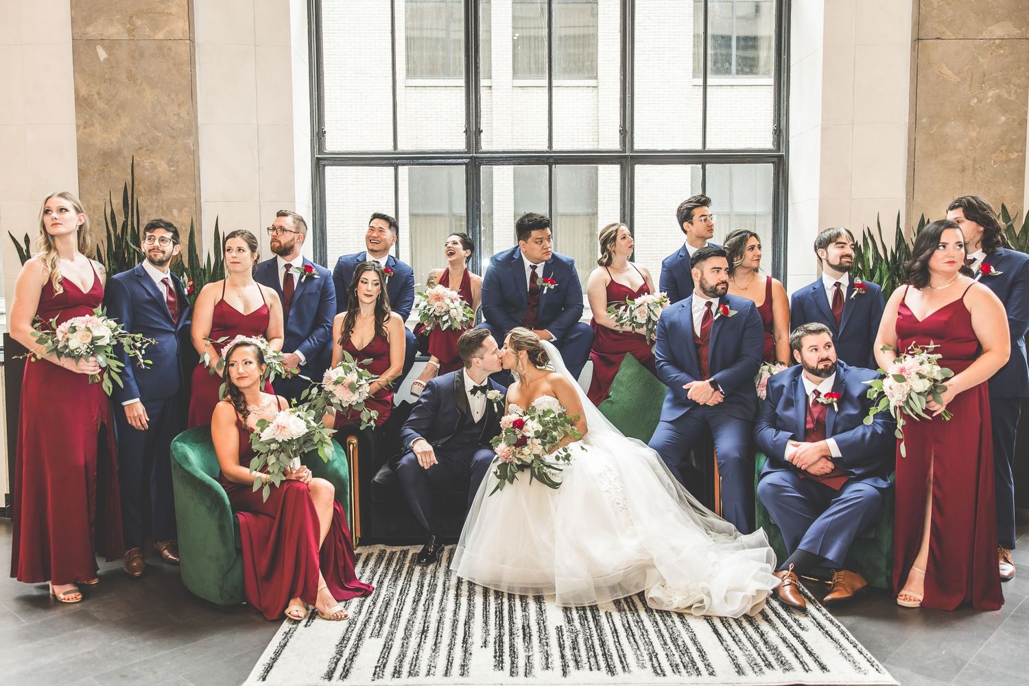 Large wedding party in burgundy and blue attire poses together in bright venue.