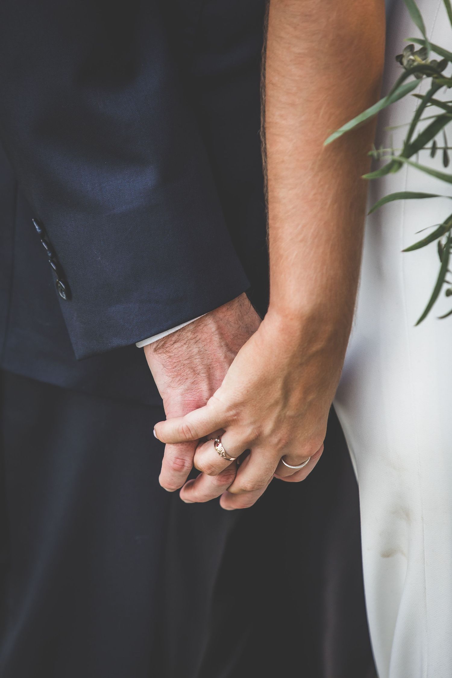 Close-up detail shot of hands with wedding rings against navy suit and white dress.