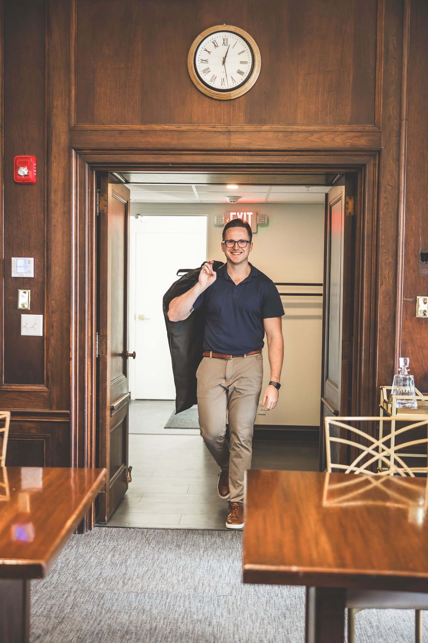 Person in navy polo and khakis carries a suit jacket while walking through wooden doorway.