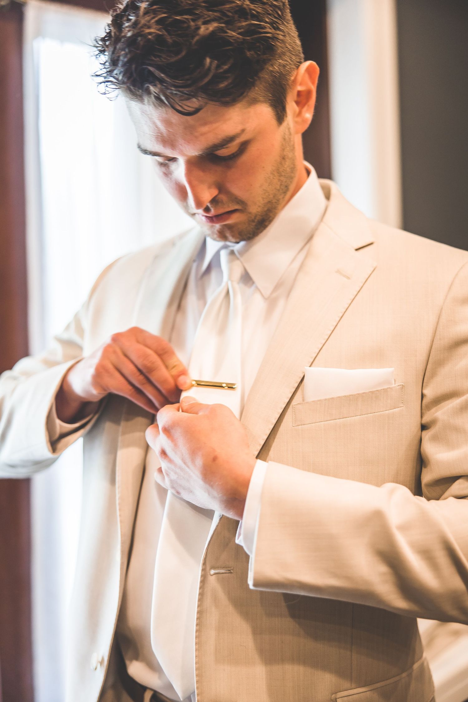 Groom in cream suit adjusts his tie while getting ready for wedding ceremony.