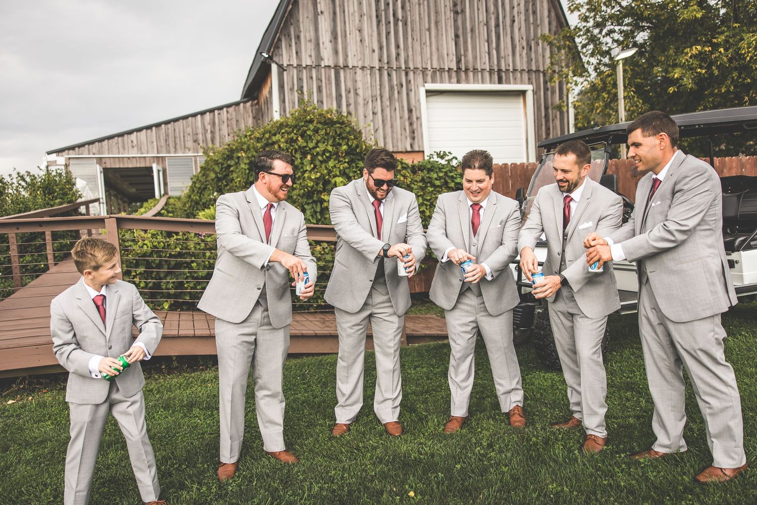 Groomsmen in light gray suits pose casually in front of a rustic barn.