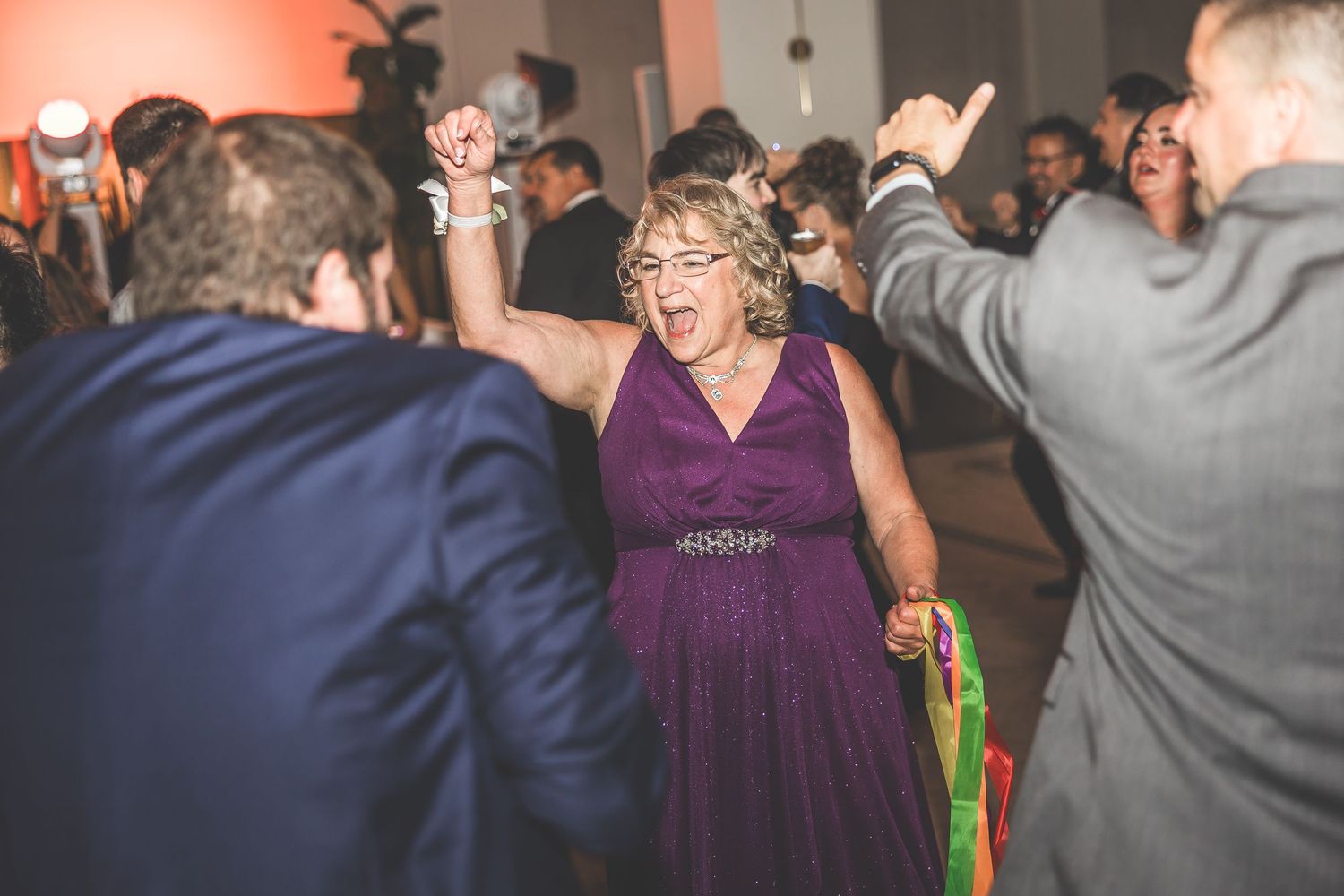 Wedding guests dance under warm lighting at an indoor reception with decorative columns.