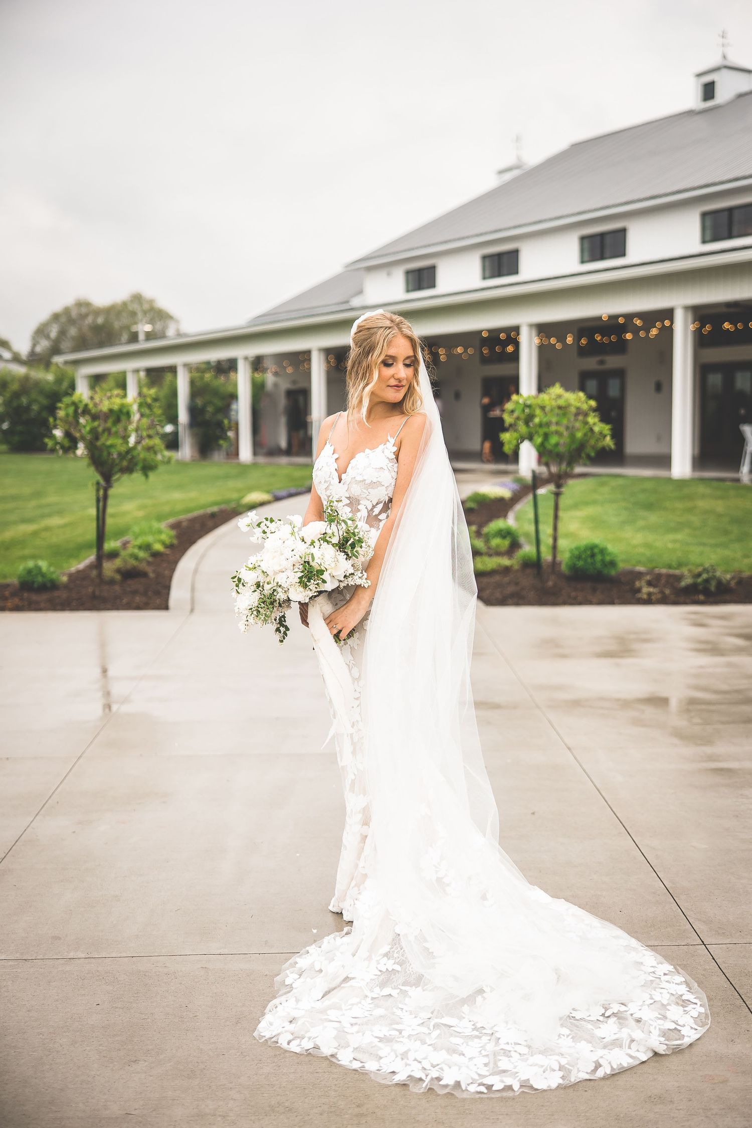 Bride in lace gown with cathedral veil poses in front of white columned venue.