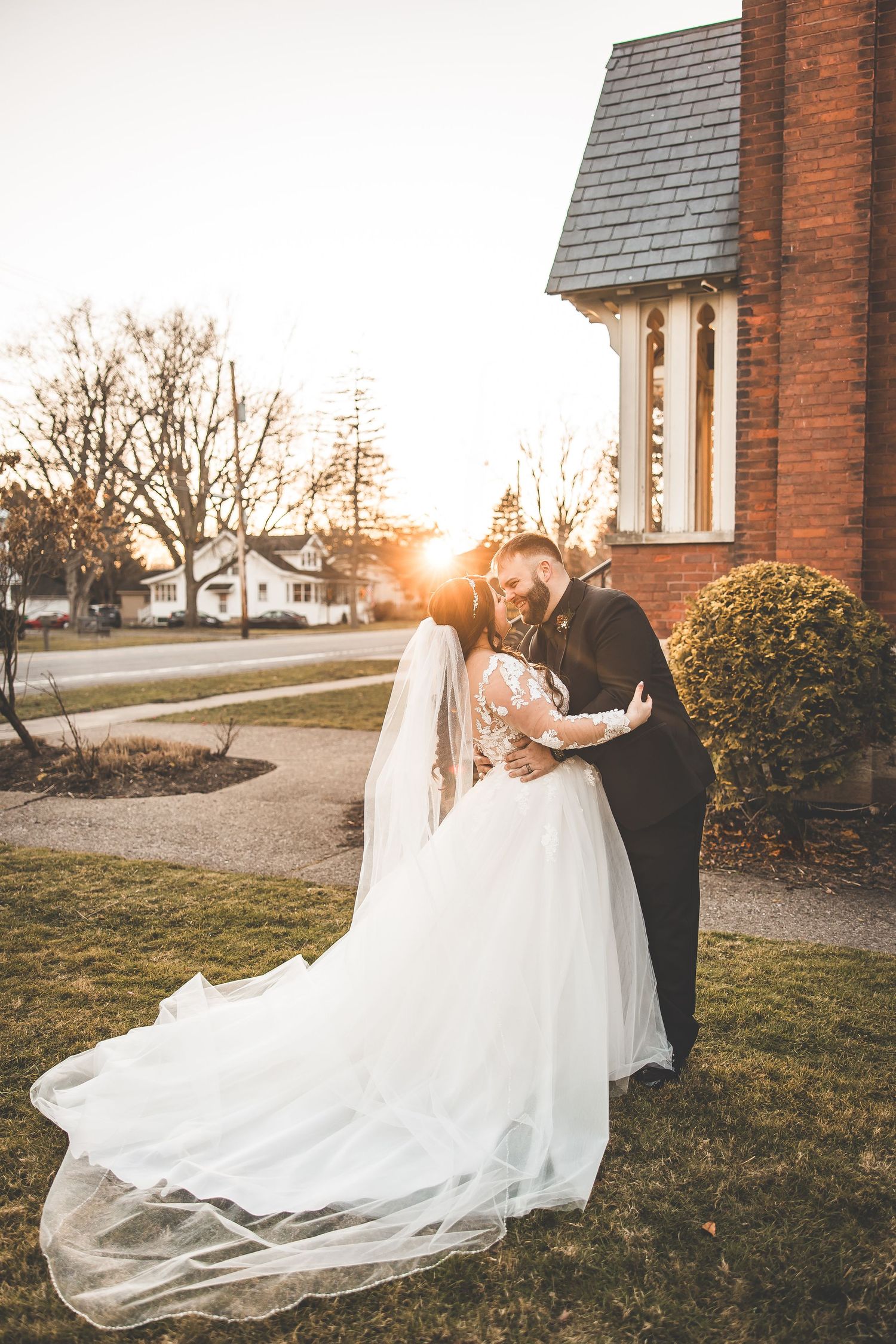 Bride's white dress flows dramatically in evening light against brick church backdrop.