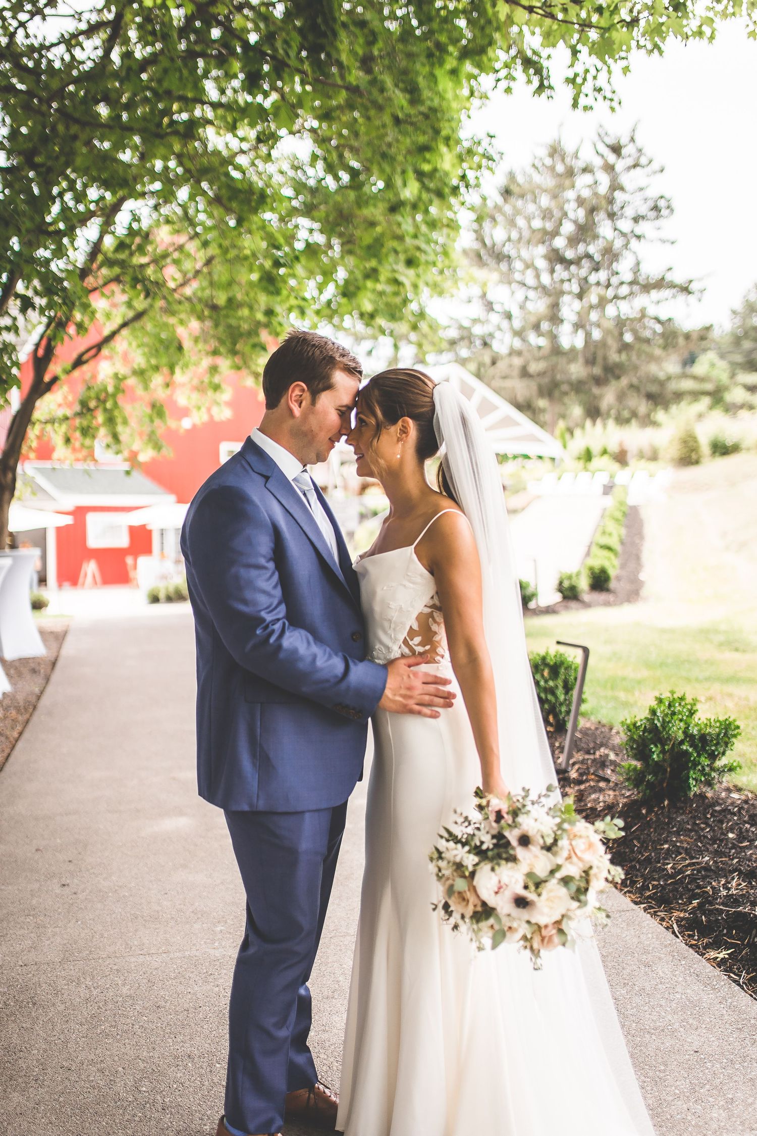 Newlyweds share a tender moment on a landscaped pathway near a red barn.