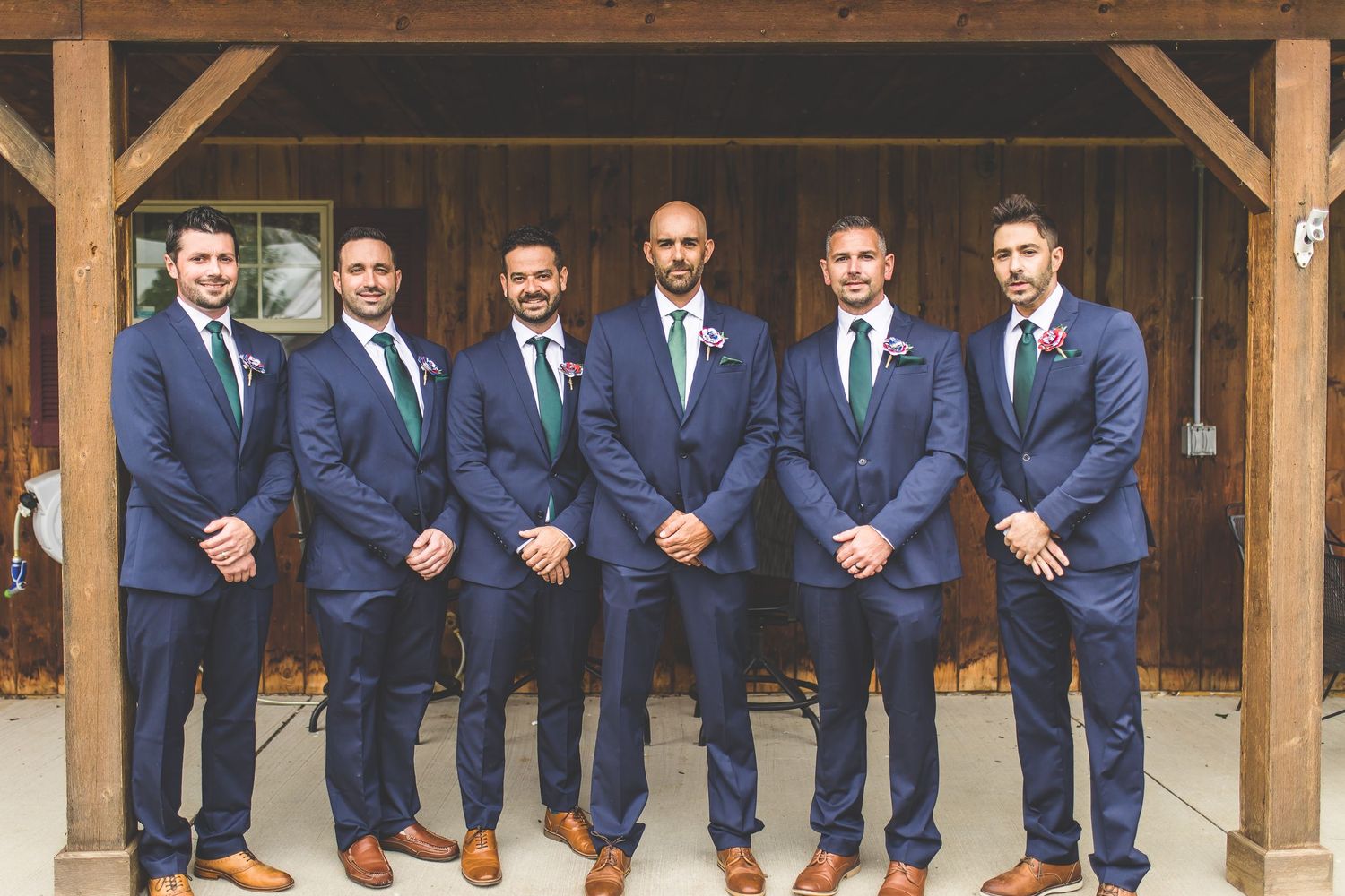 Wedding party in navy suits and green ties stand in line under wooden beams.