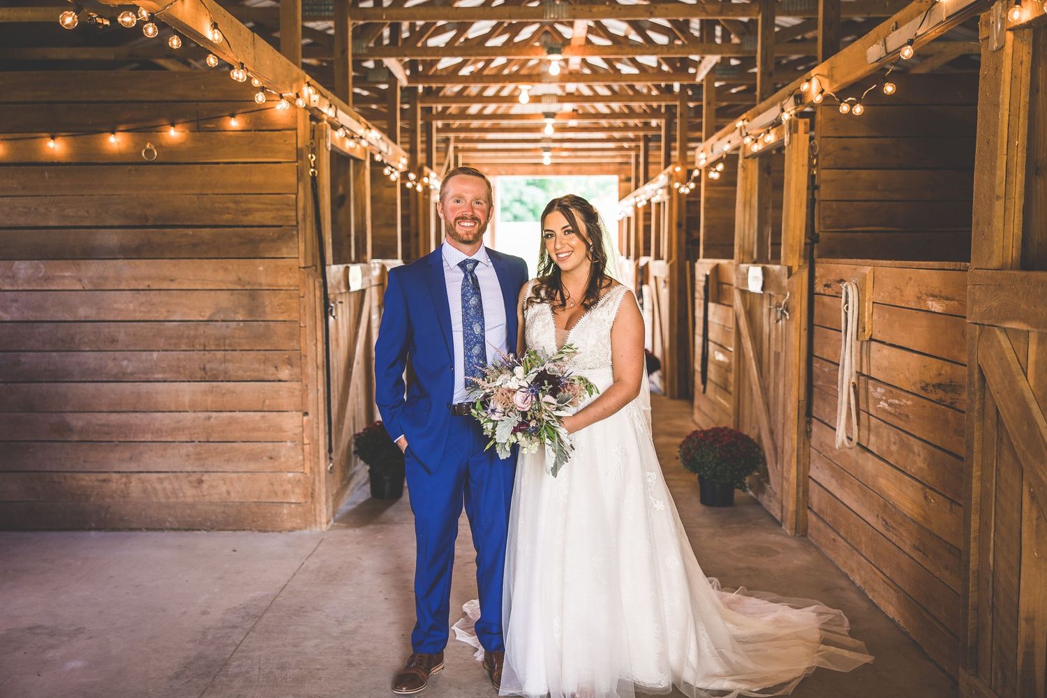 Wedding couple poses in rustic barn venue with string lights creating warm ambiance.