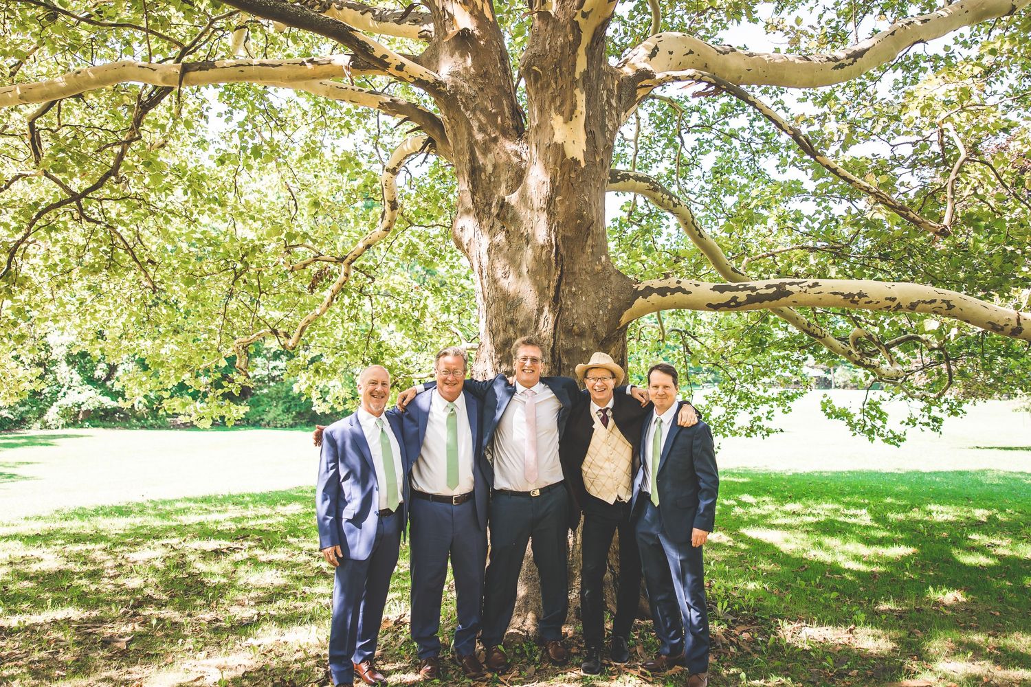Groomsmen pose playfully under a large tree branch in a garden setting.