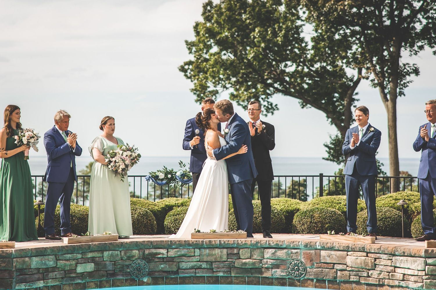 Wedding party lined up along stone wall with green trees and blue sky in background.