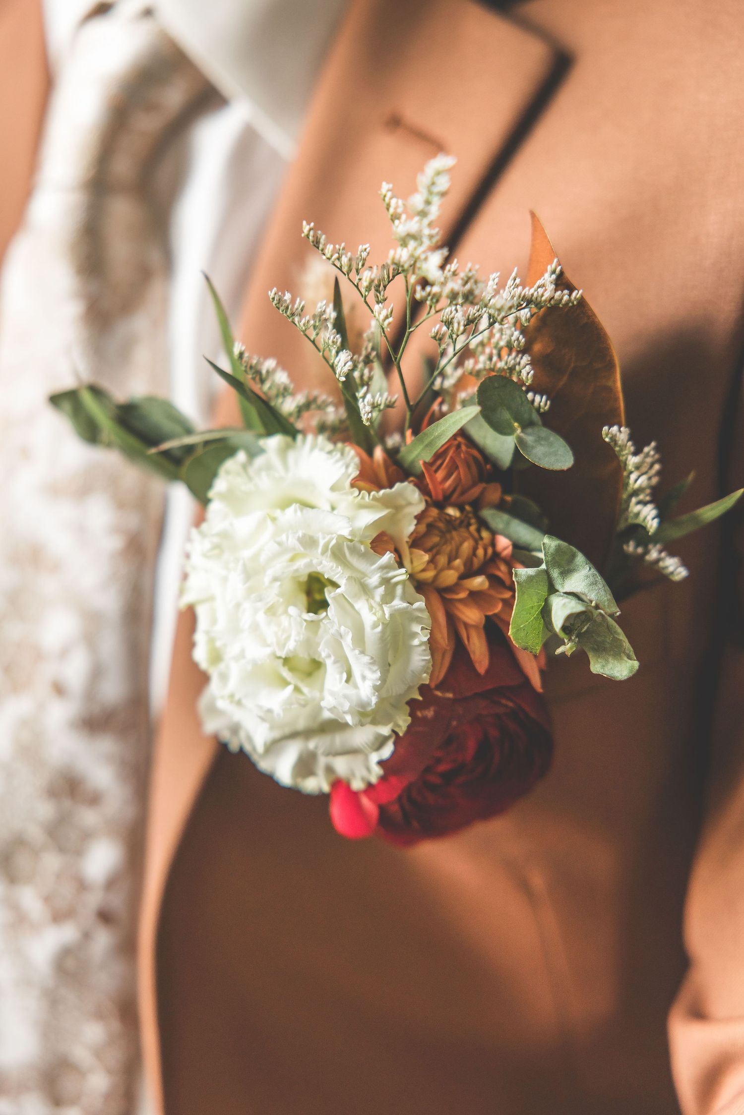 Close-up of a wedding boutonniere with white flowers and greenery against a brown background.