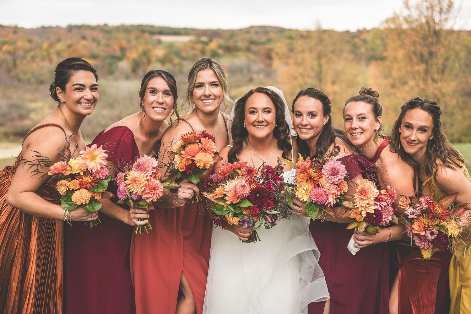 Wedding party in burgundy and mustard dresses poses with colorful fall bouquets outdoors.