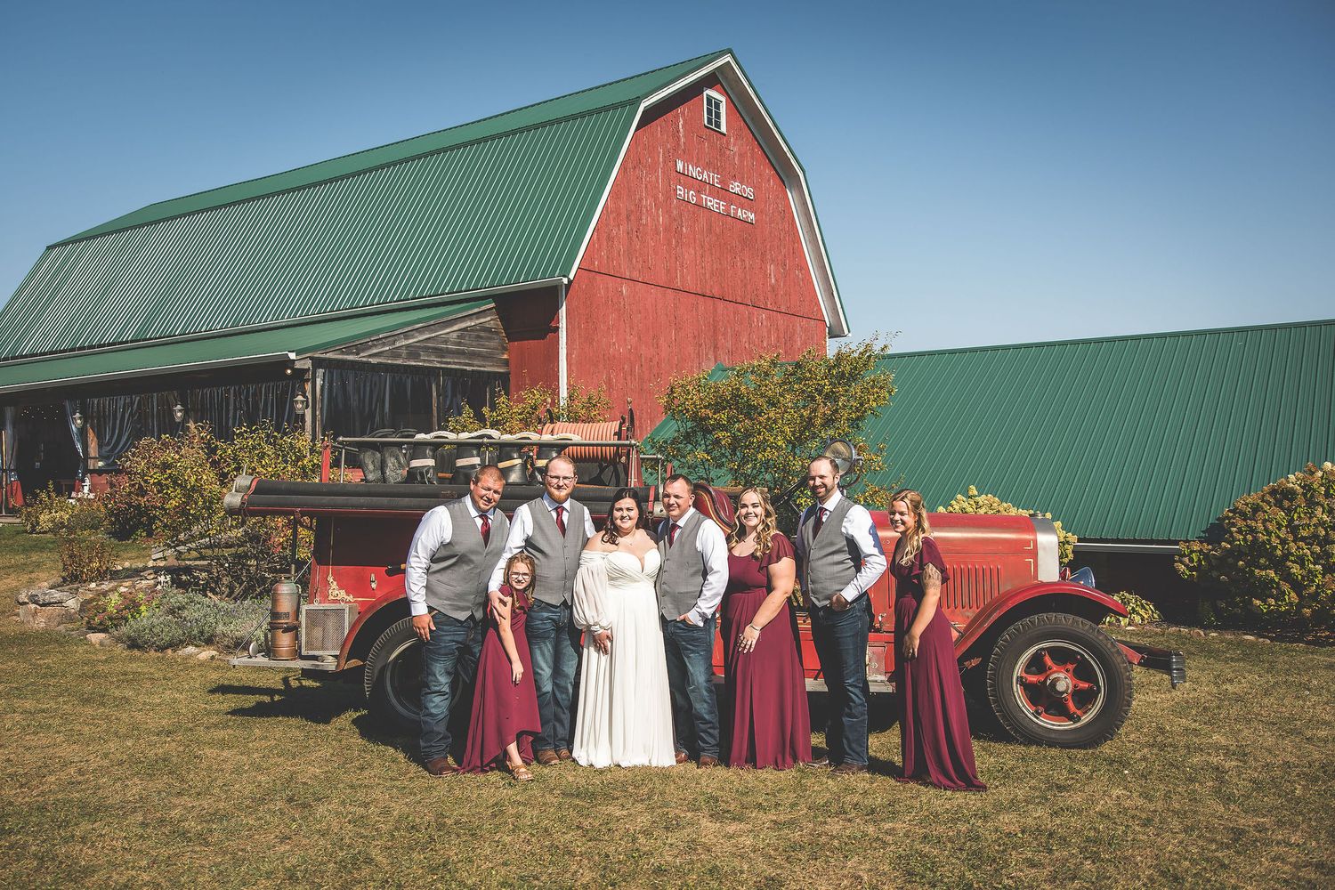 Wedding party gathers for group photos in front of red barn on sunny autumn day.