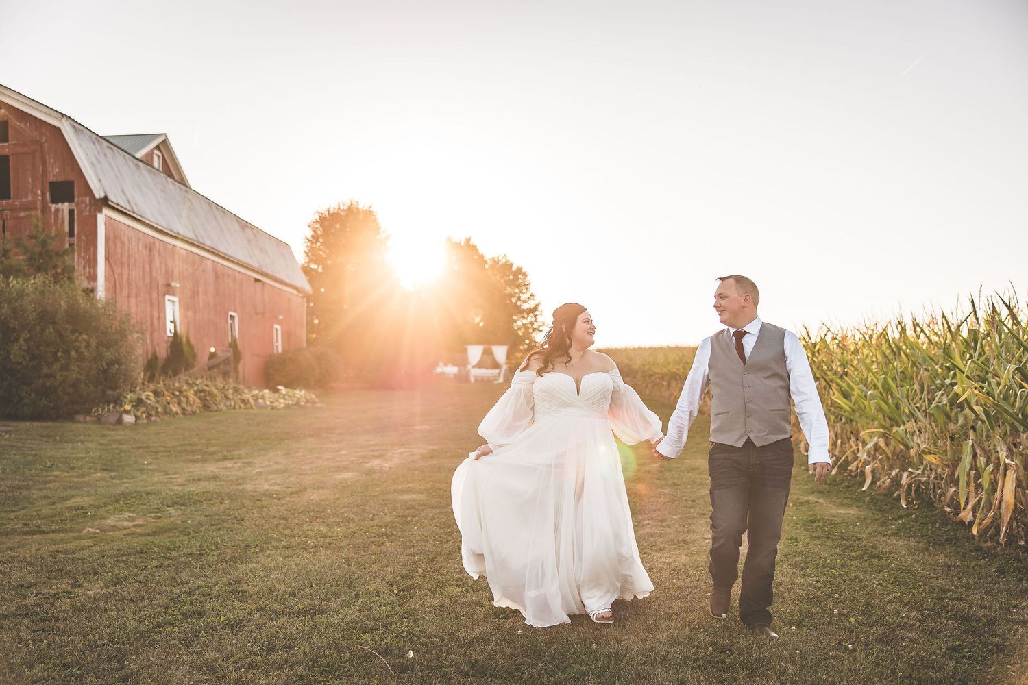 Newlyweds walk hand in hand through a field at golden hour with a barn in the background.