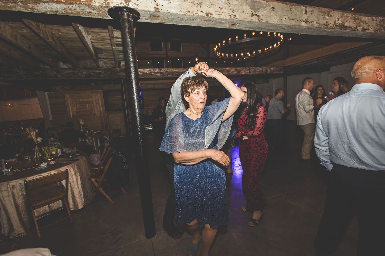Wedding guests dance and celebrate in a rustic barn venue with string lights overhead.