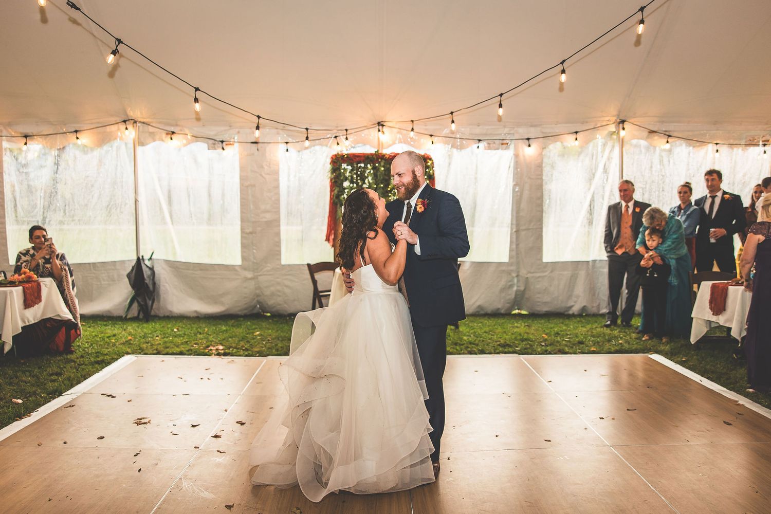 Wedding reception dance floor illuminated by string lights in a white tent with guests watching.
