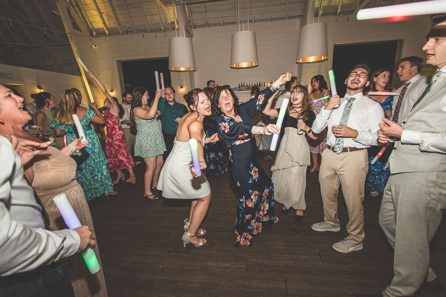 Wedding guests dance while holding glowing foam sticks at an indoor reception.