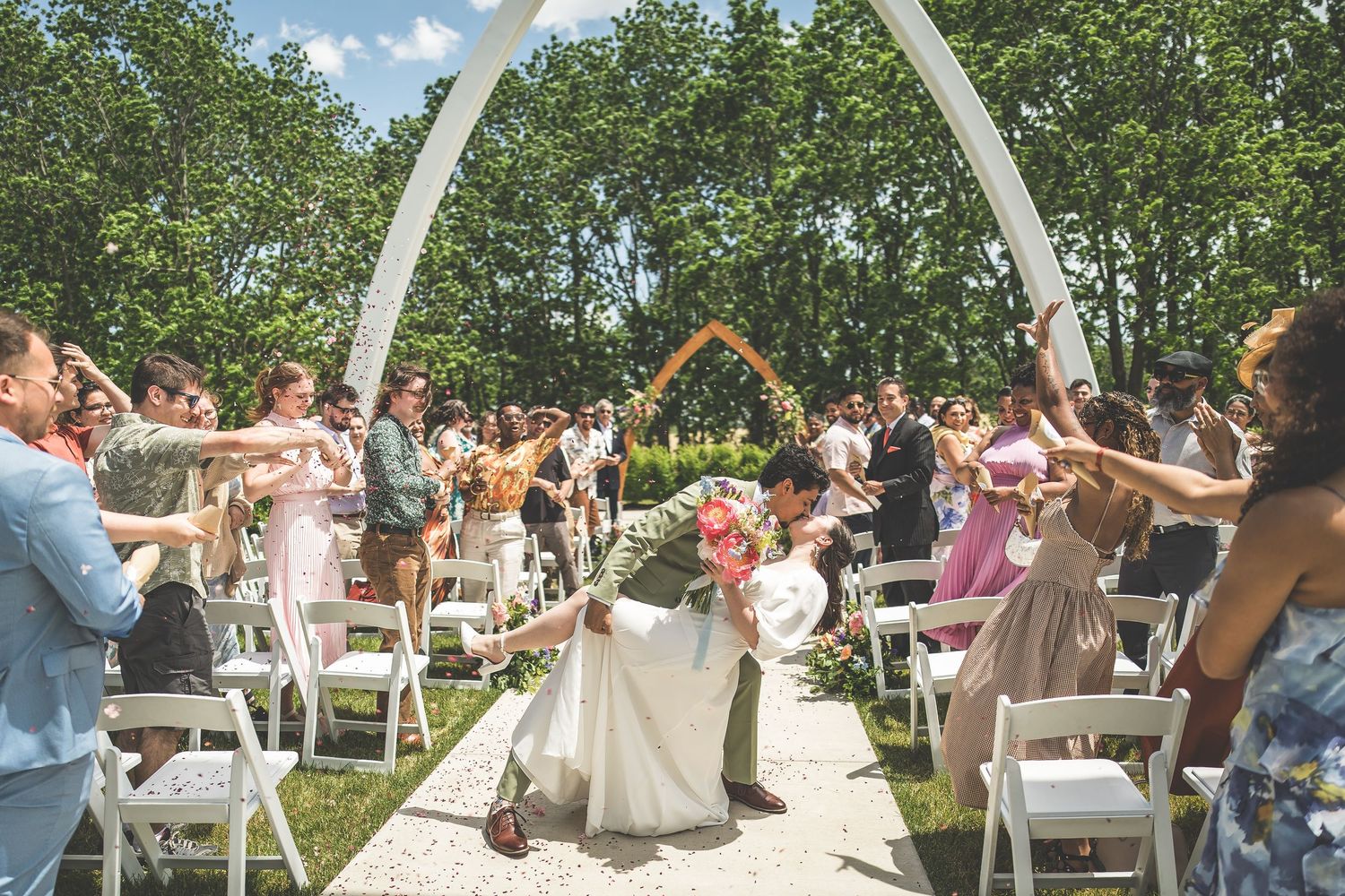 Bride and groom share a romantic dip kiss while guests cheer along a flower-adorned arch aisle at an outdoor wedding.