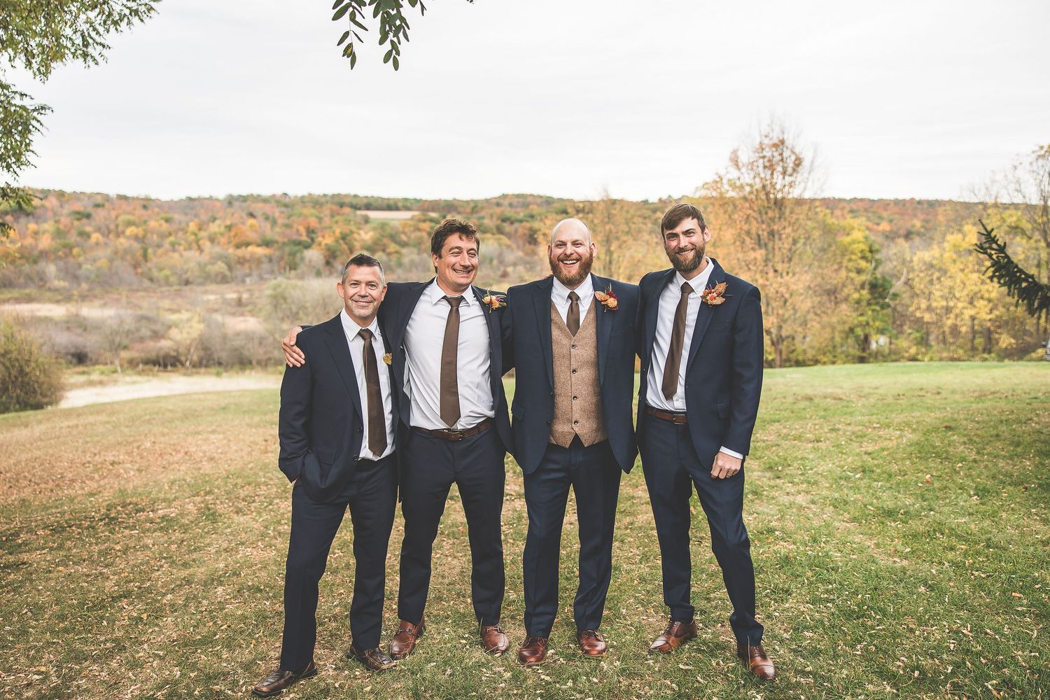 Groomsmen in dark suits pose together on rolling autumn hillside.