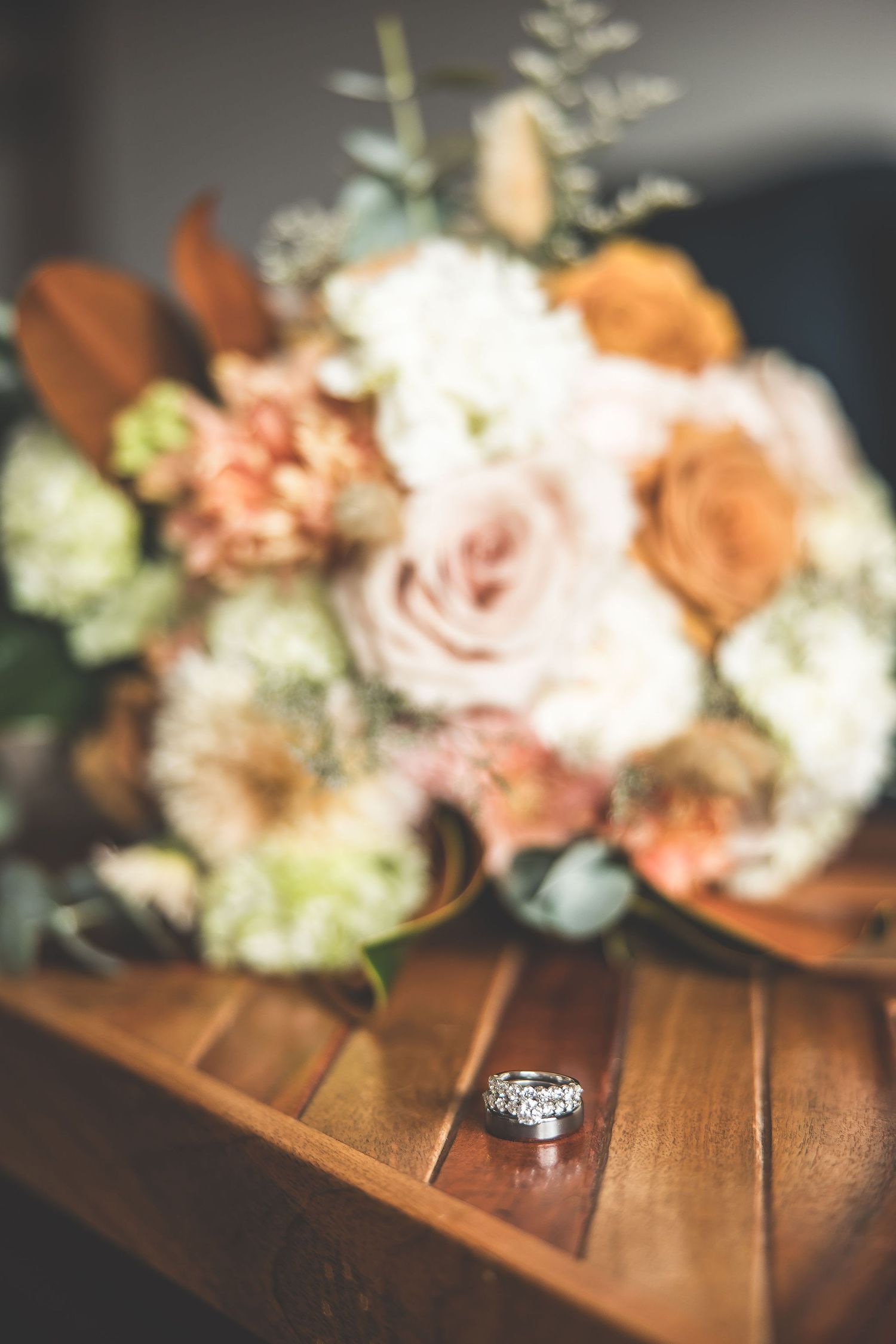Wedding rings photographed with soft focus peach and white floral arrangements on wooden surface.