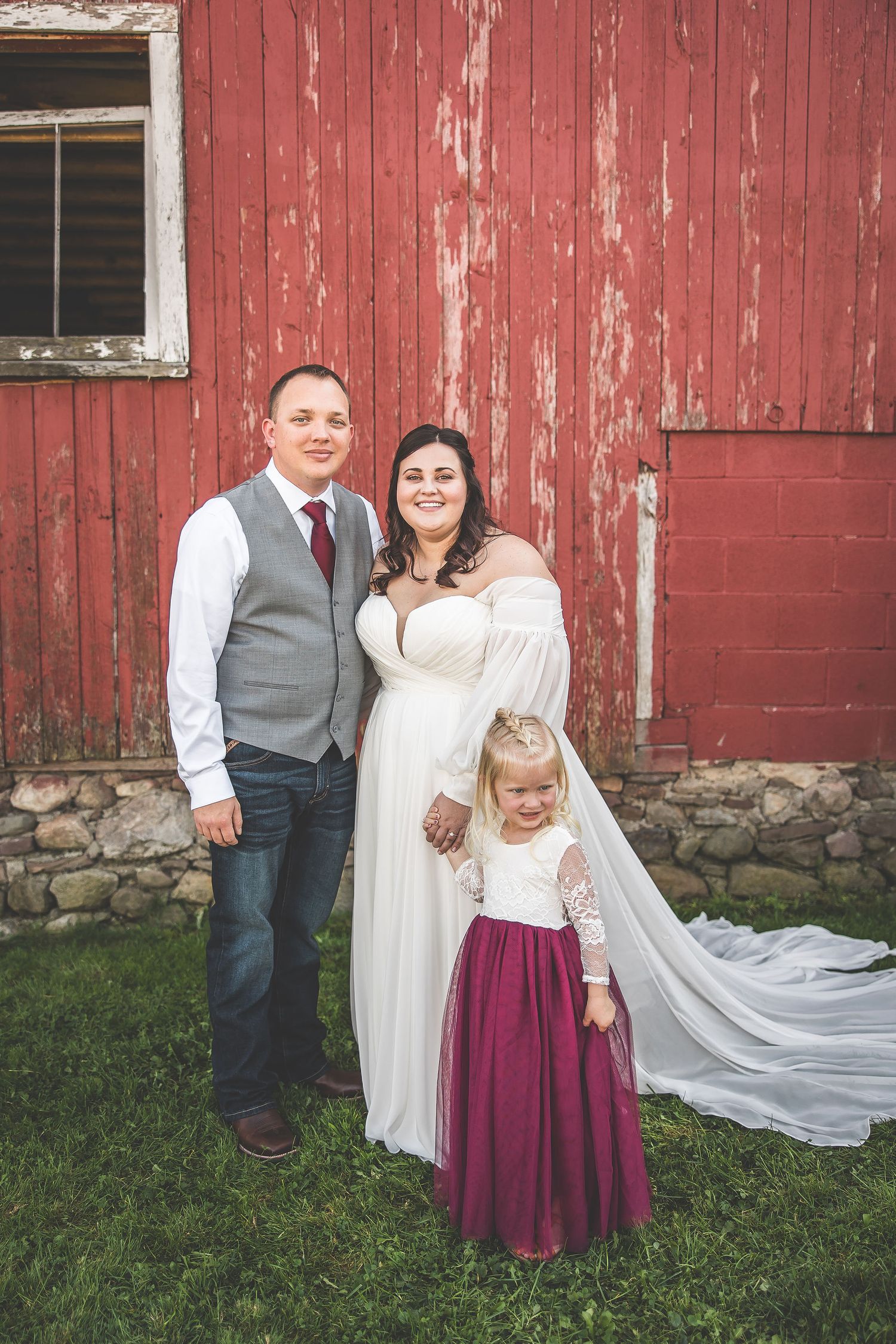 Wedding party poses in front of a classic red barn with flowing white dress details and burgundy accents.
