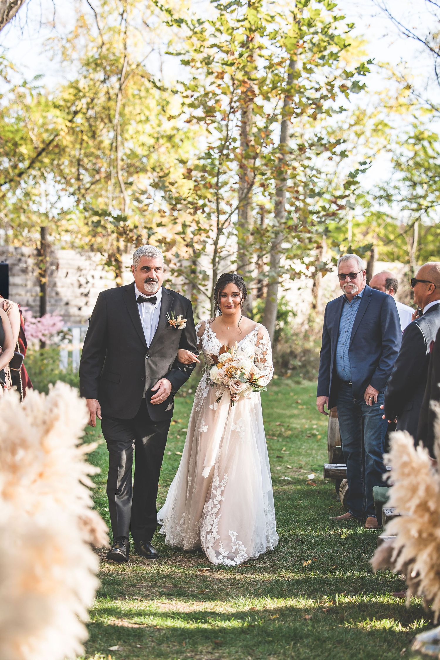 Wedding guests gather on a grassy lawn surrounded by autumn trees during an outdoor ceremony.