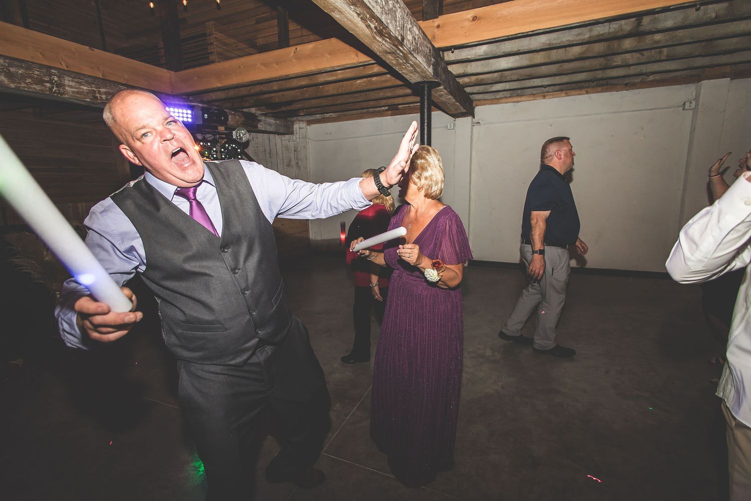 Wedding guests dance and celebrate in a rustic barn venue with wooden beams and ambient lighting.