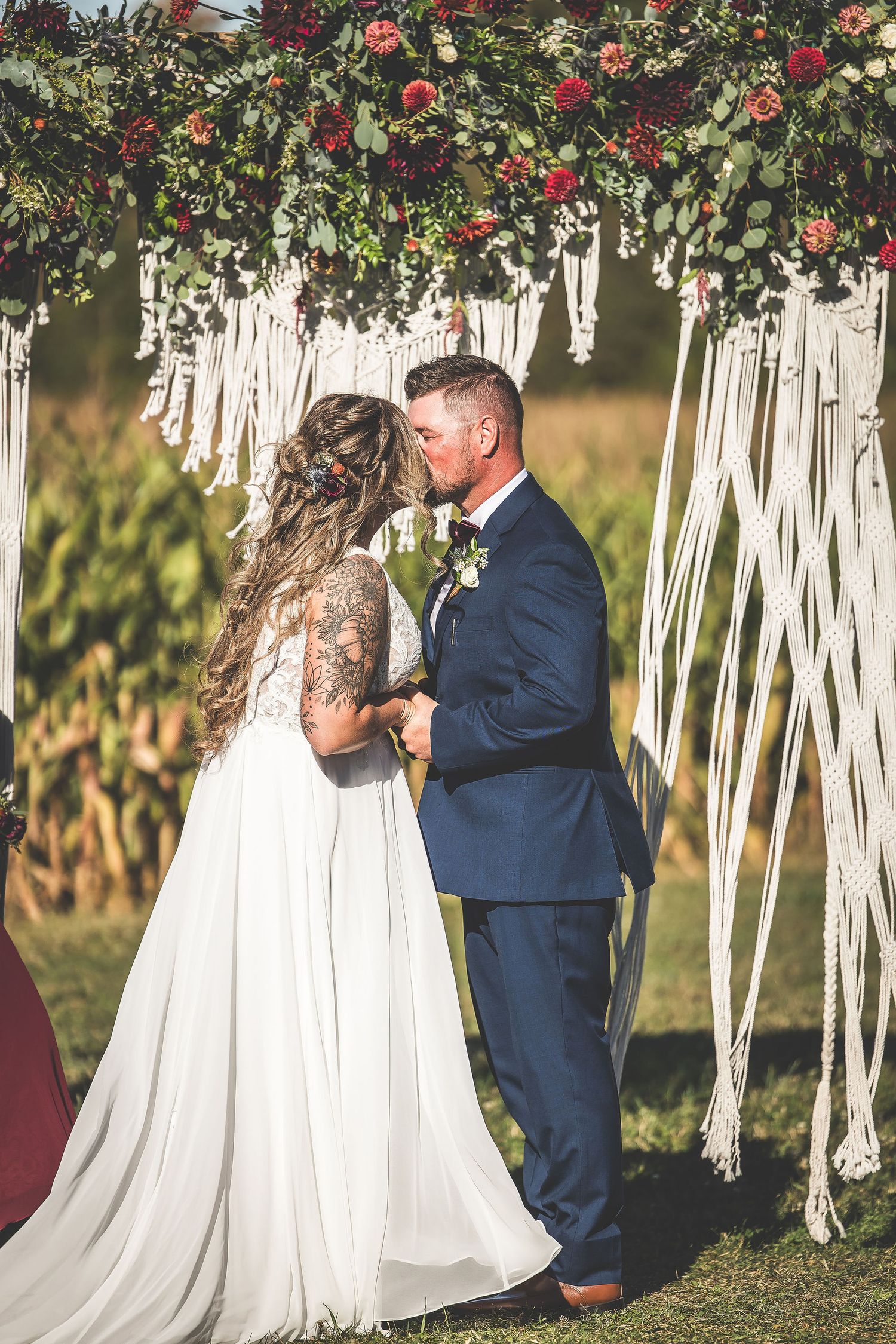 Intimate outdoor wedding ceremony with white macrame backdrop and red floral accents.