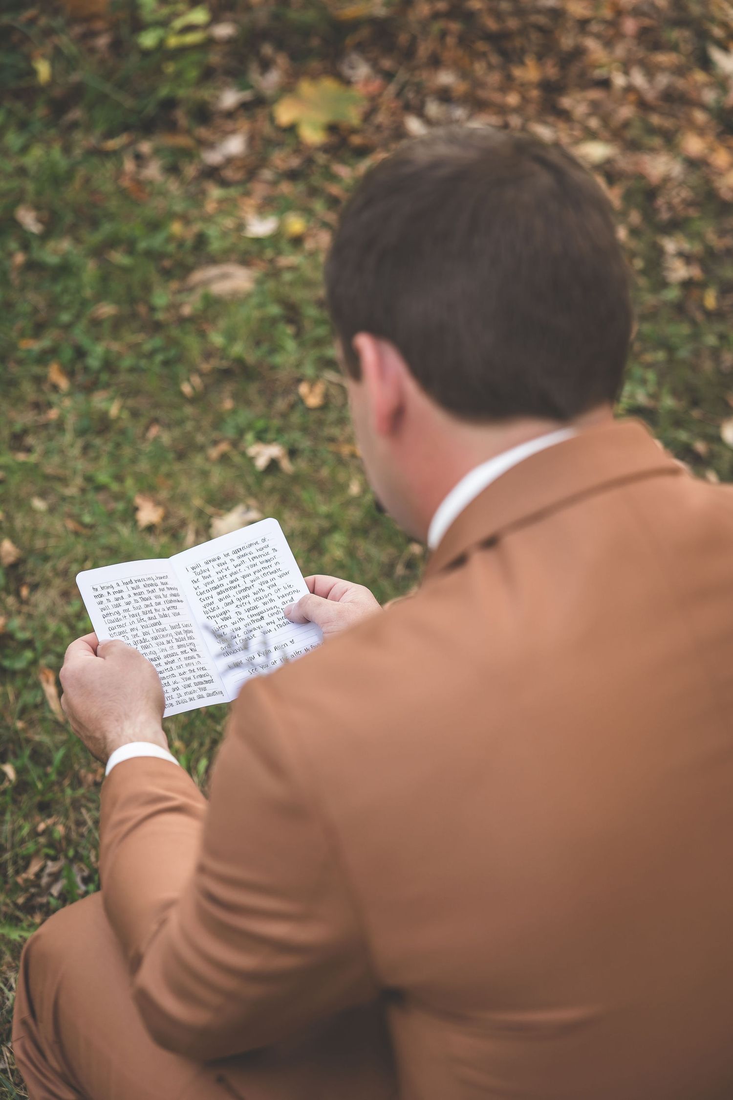A person in a brown suit jacket reads from a small book while sitting outdoors among fallen autumn leaves.