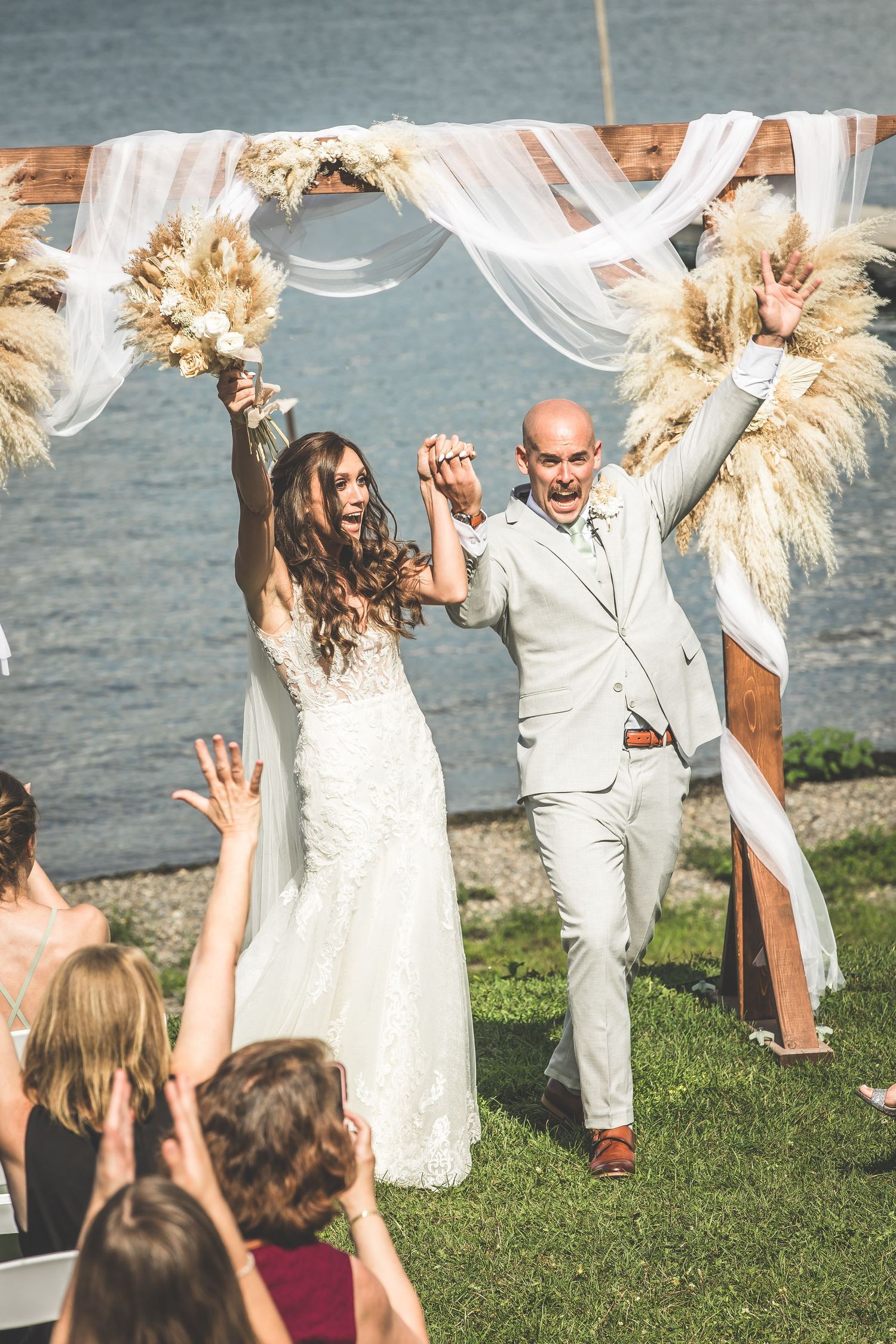Newlyweds celebrate with guests on a sunny lakeside lawn during their outdoor wedding reception.