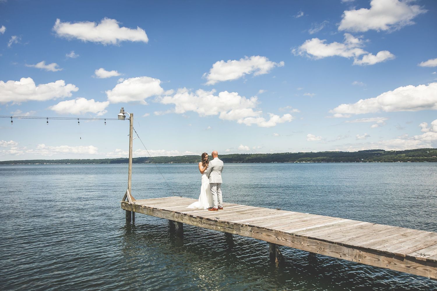A distant view of newlyweds embracing on a long wooden pier over calm blue waters.