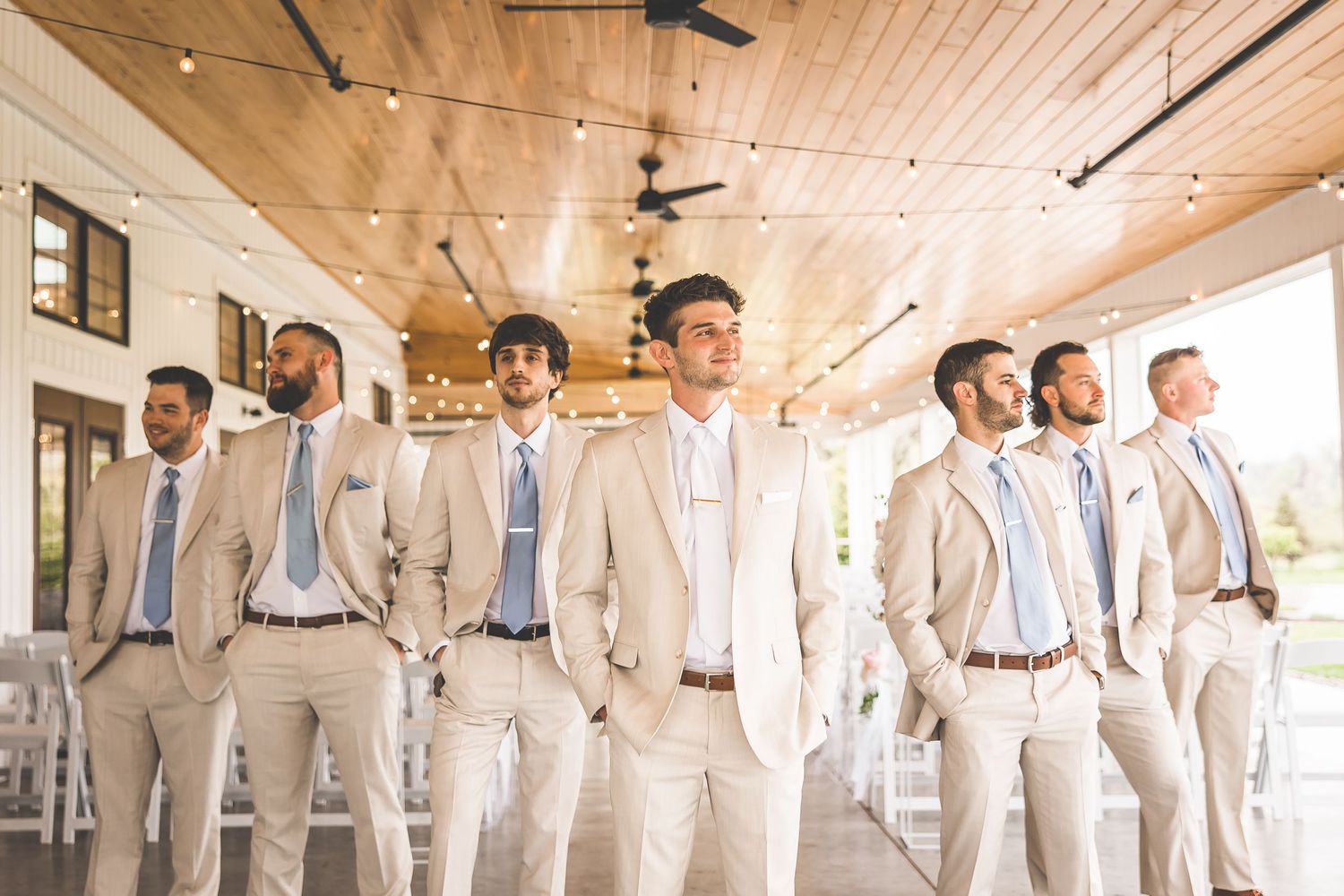 Wedding party in light beige suits standing in line at rustic indoor venue.