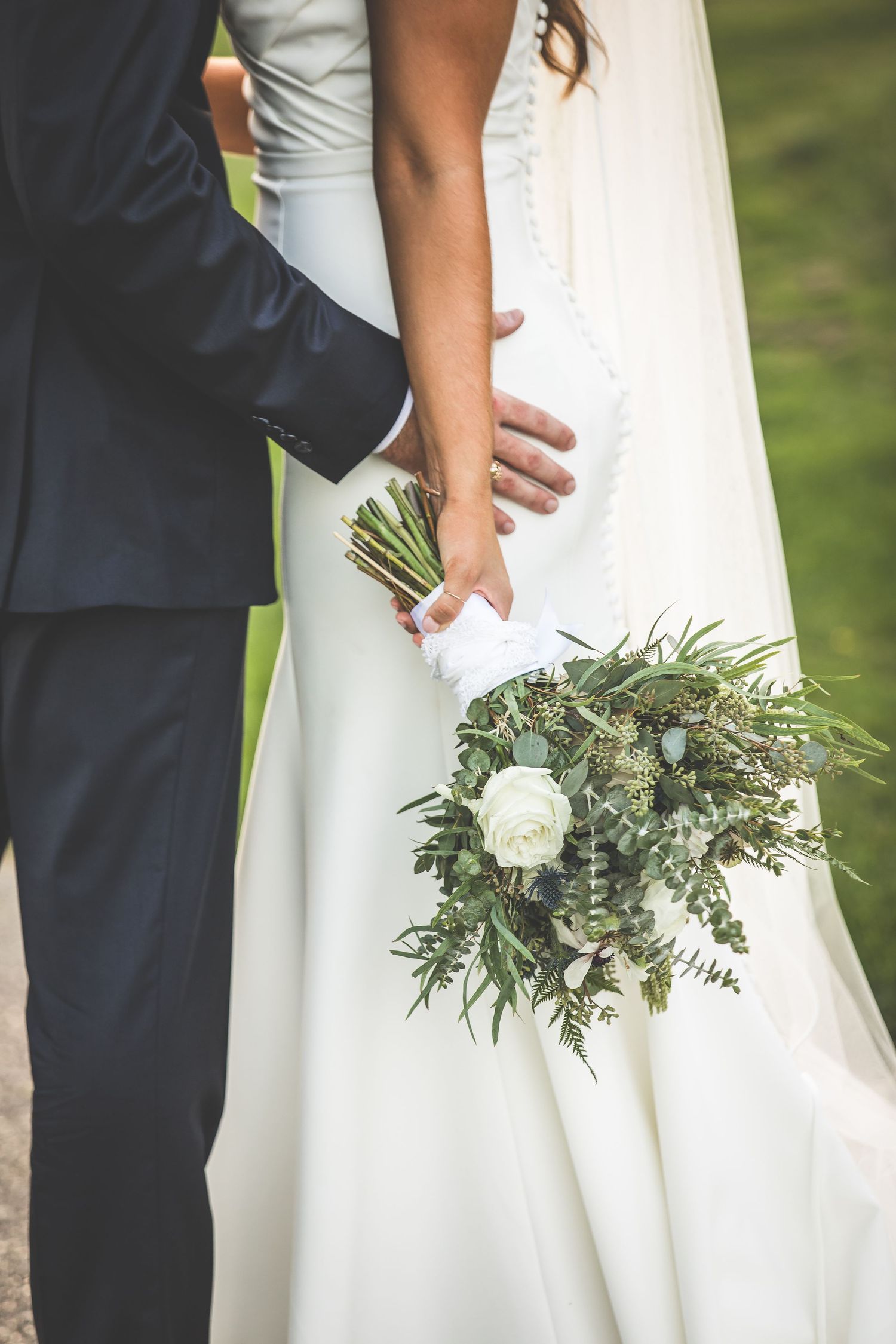 Detail shot of white bridal bouquet with greenery against wedding dress and navy suit.
