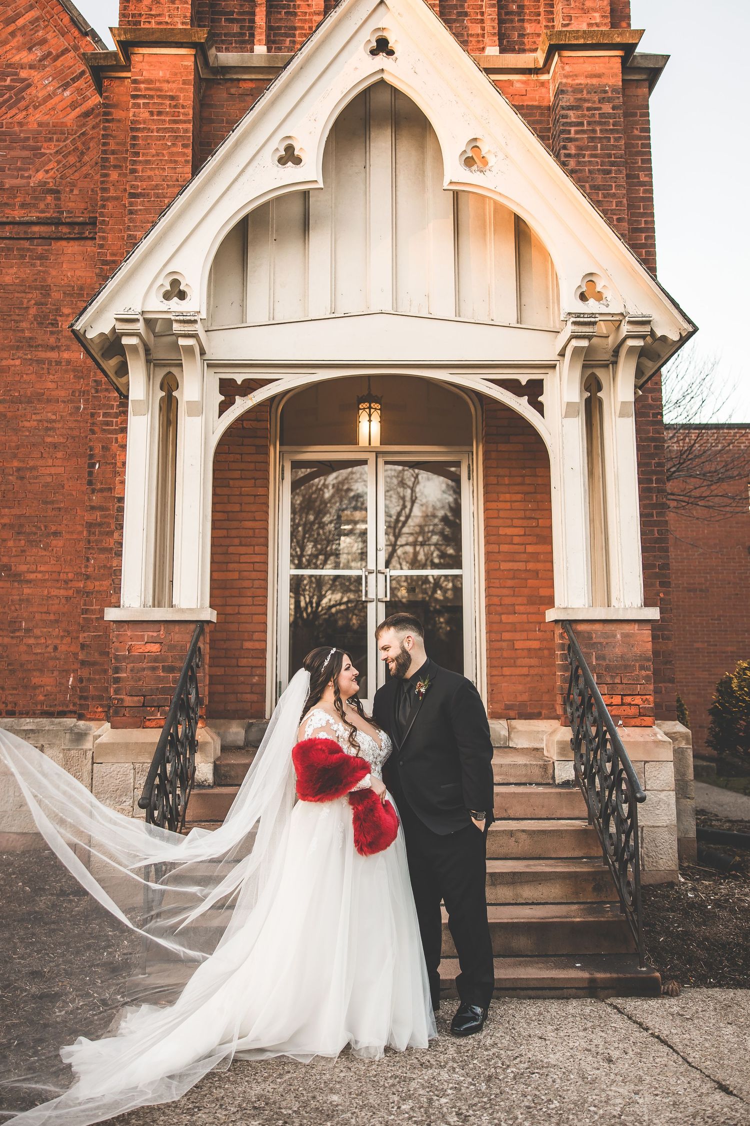 Bride in white gown with red accent poses under gothic church archway.